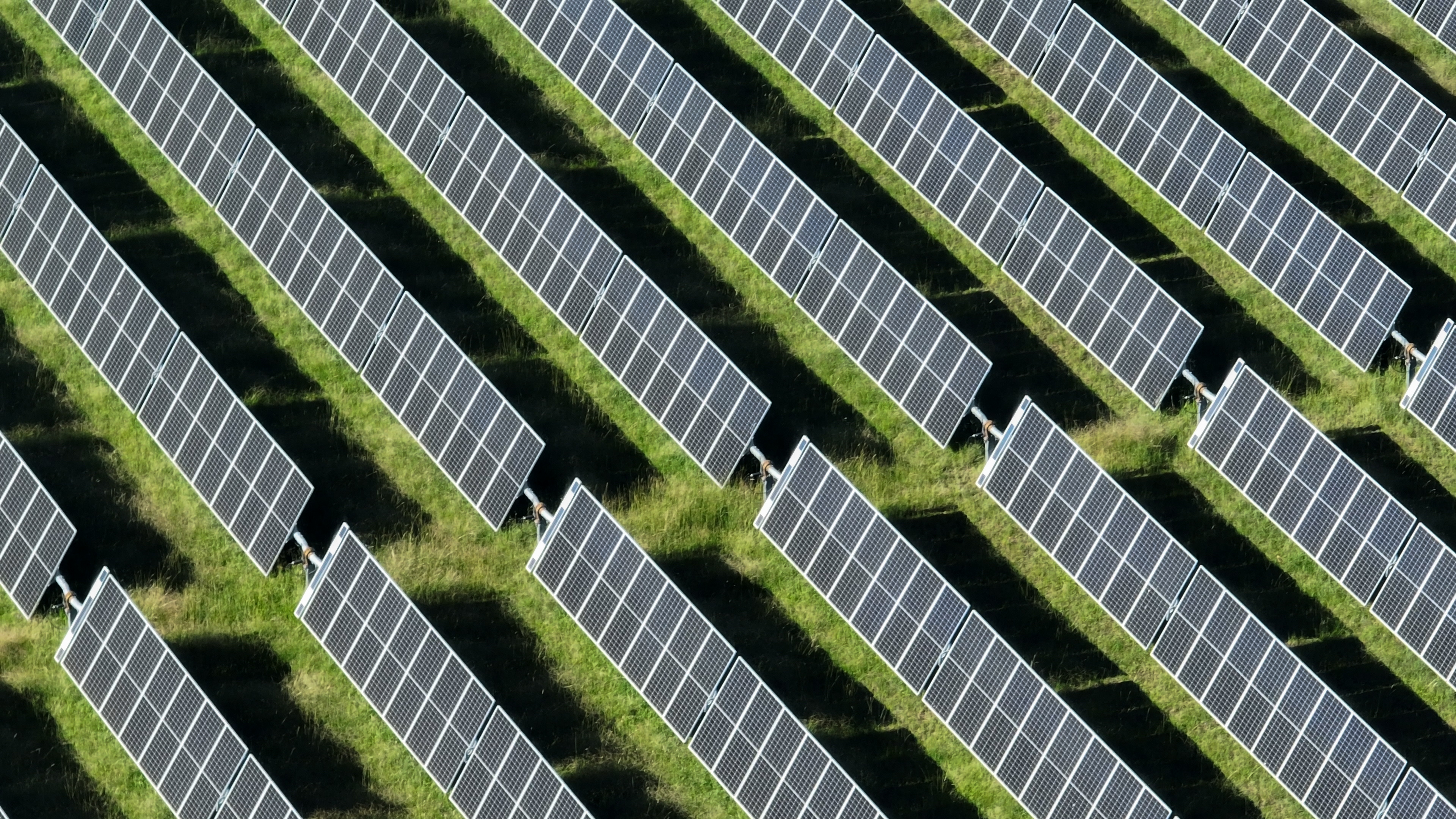 This stunning aerial view captures an  array of solar panels arranged in neat, parallel rows across the landscape. From above, the panels shimmer under the bright sunlight, creating a striking contrast against the natural terrain below. The organized rows of solar panels stretch across acres of land, symbolizing the growing global shift toward renewable energy. The grid-like pattern highlights the efficiency and scale of modern solar farms, contributing to sustainable energy production.This high-resolution image showcases the incredible reach and potential of solar power as a clean, renewable energy source. Whether situated in rural fields, expansive deserts, or atop rooftops, these solar panels represent a major step toward reducing carbon footprints and combating climate change. The solar farm's orderly rows and reflective surfaces create a visually appealing scene, demonstrating both technological innovation and environmental responsibility.