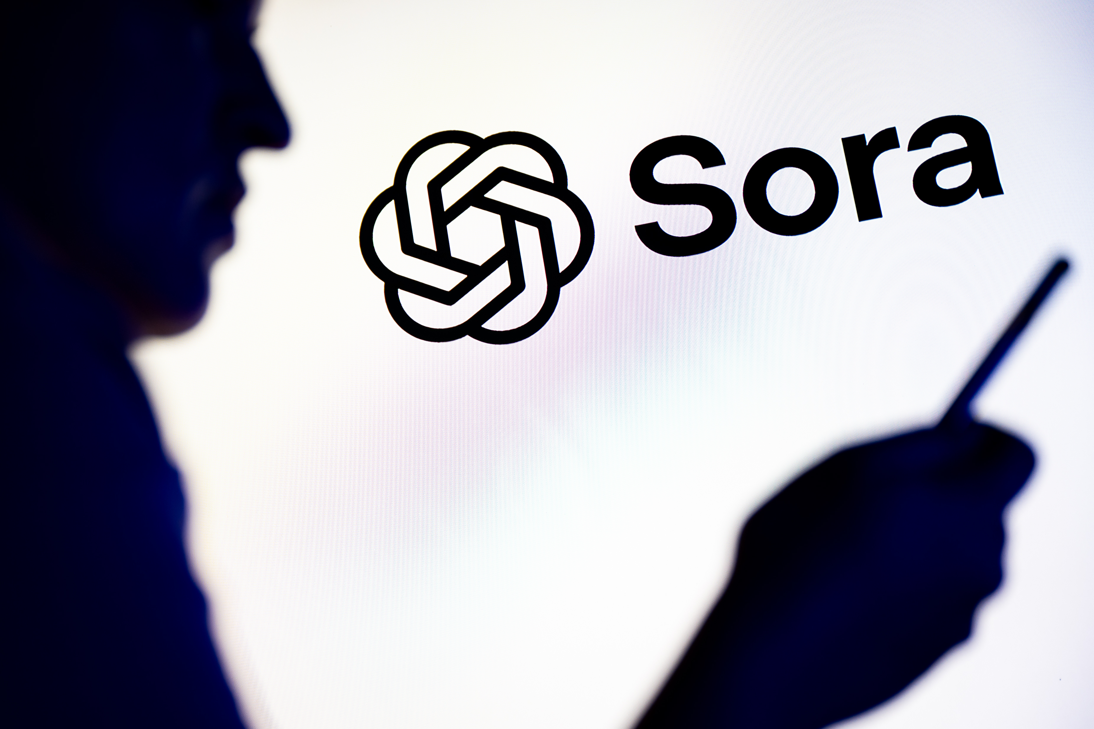 POLAND - 2025/11/26: In this photo illustration, a silhouetted individual is seen holding a mobile phone with a Sora of ChatGPT OpenAI logo displayed in the background. (Photo Illustration by Mateusz Slodkowski/SOPA Images/LightRocket via Getty Images)