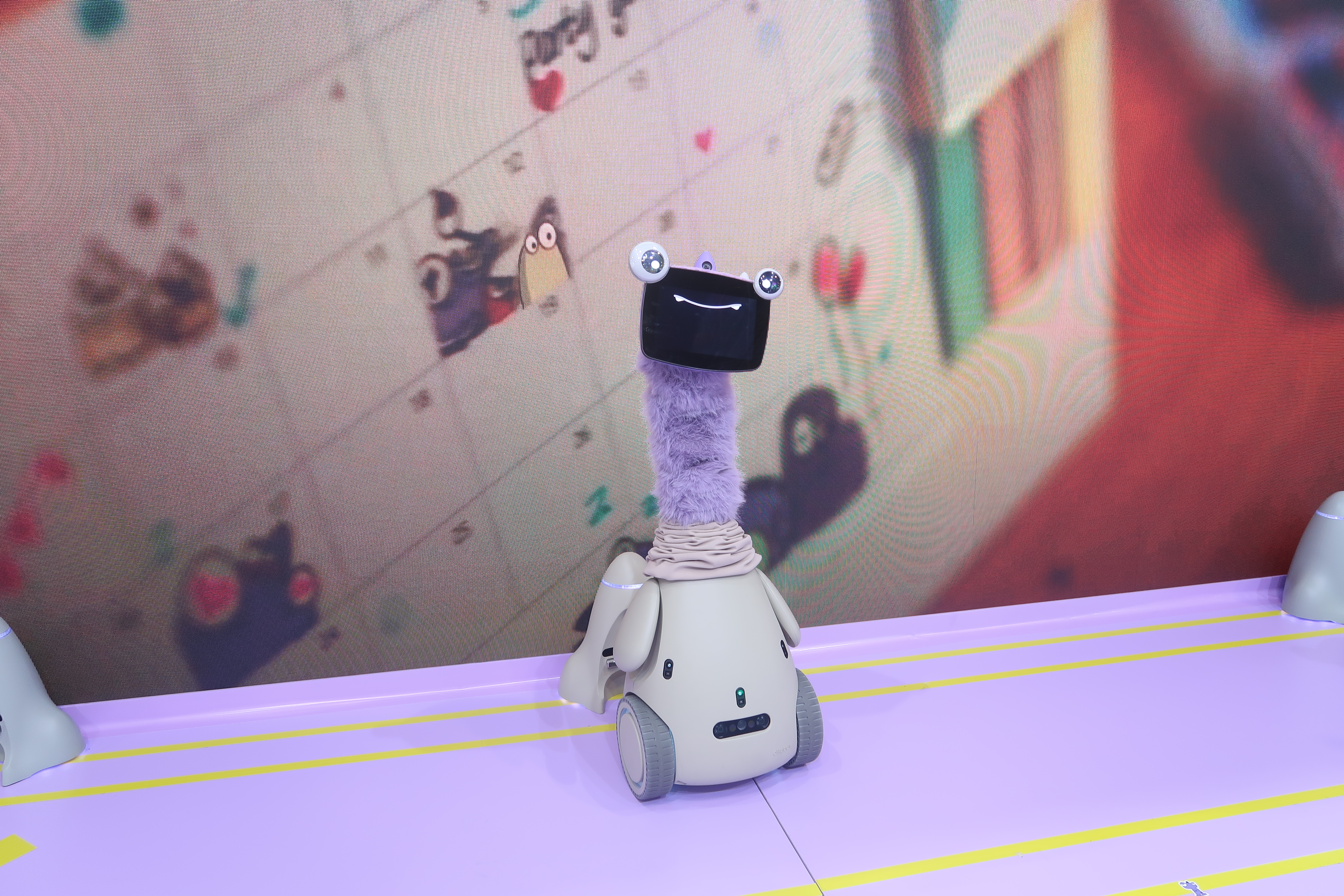 An OlloBot robot with a long purple neck stands in front of a display showing a home calendar covered in stickers
