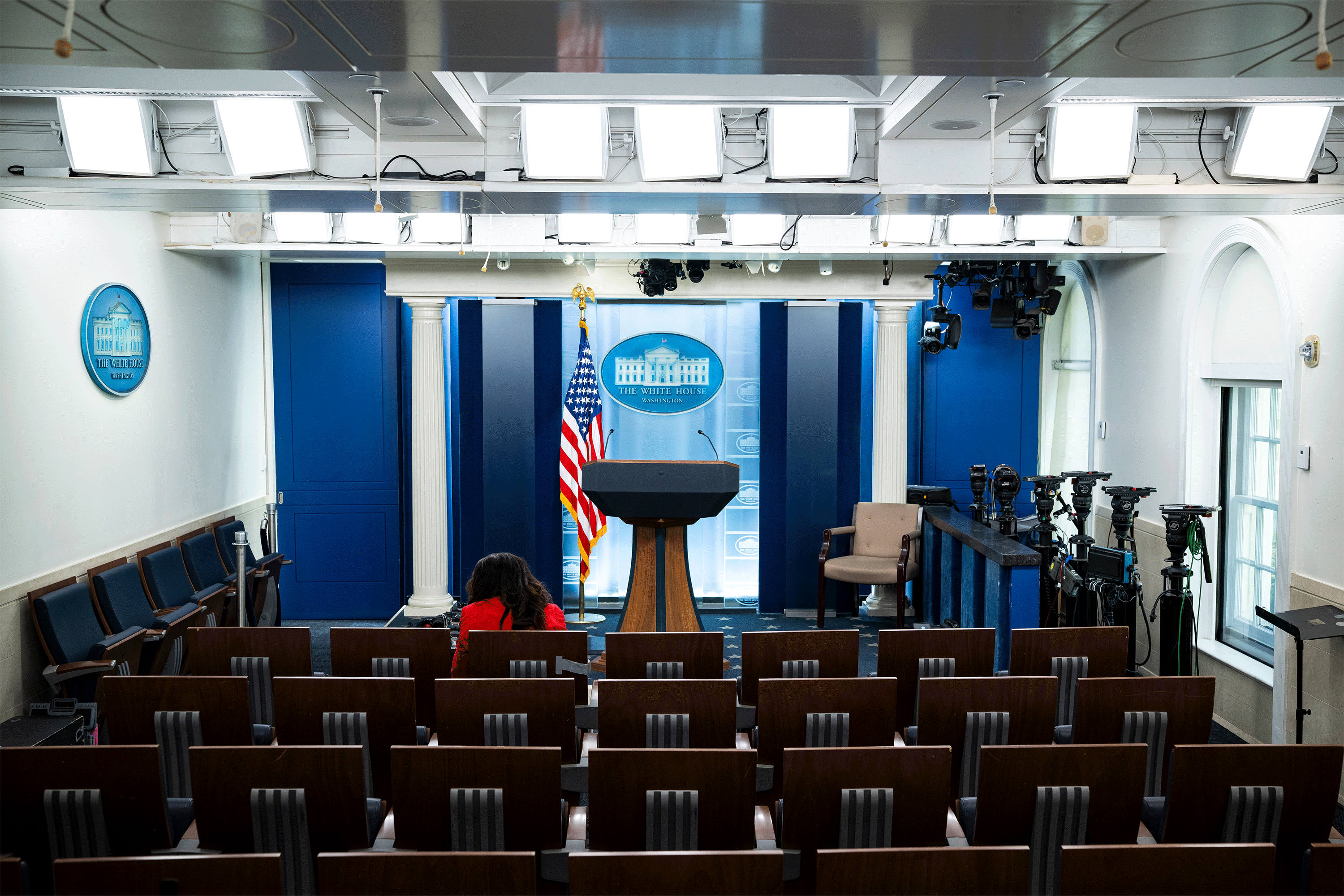 The White House Briefing Room is empty except for one reporter sitting in the front row wearing a red blazer and facing away from the camera.